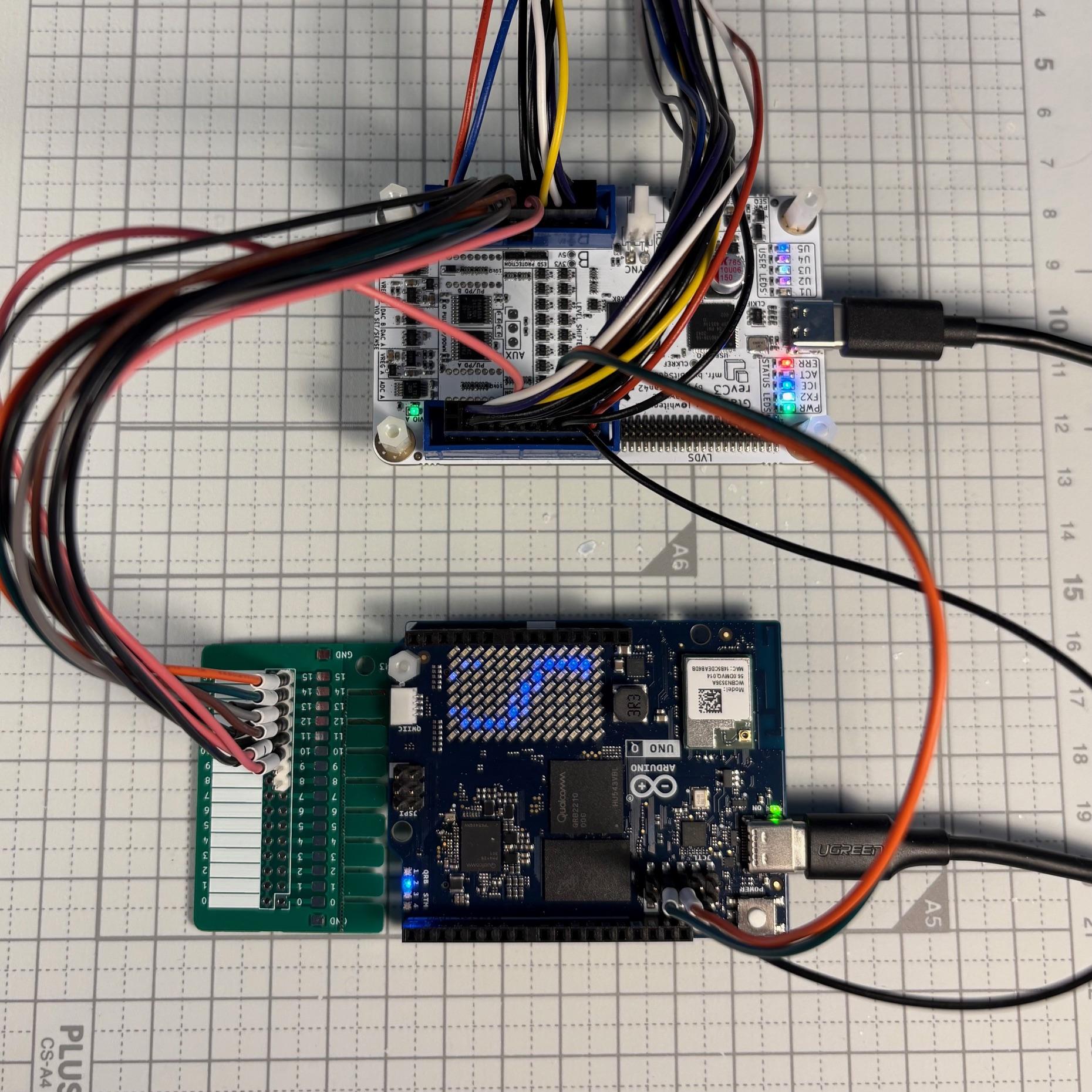 A glasgow board connected to an Arduino Uno Q board that has a little pin header breakout PCB attached to some testpoints. Both boards are powered up and have several LEDs lit.