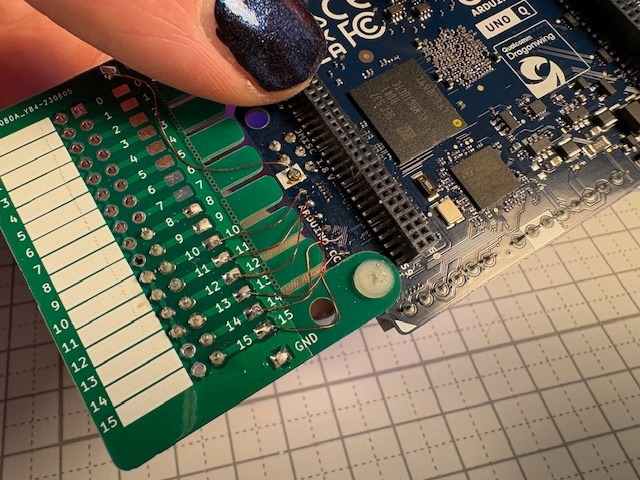 The back side of the Uno Q shown in the header picture. Thin copper wires are visible that connec the tiny testpoints at the board edge to a green breakout PCB. The breakout PCB has two rows of pin headers with empty white label fields next to them. The pins are numbered.
